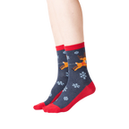 Lifestyle shot showing the socks on a person's feet. The socks are dark gray with red cuffs and toes, featuring a reindeer and snowflake pattern.