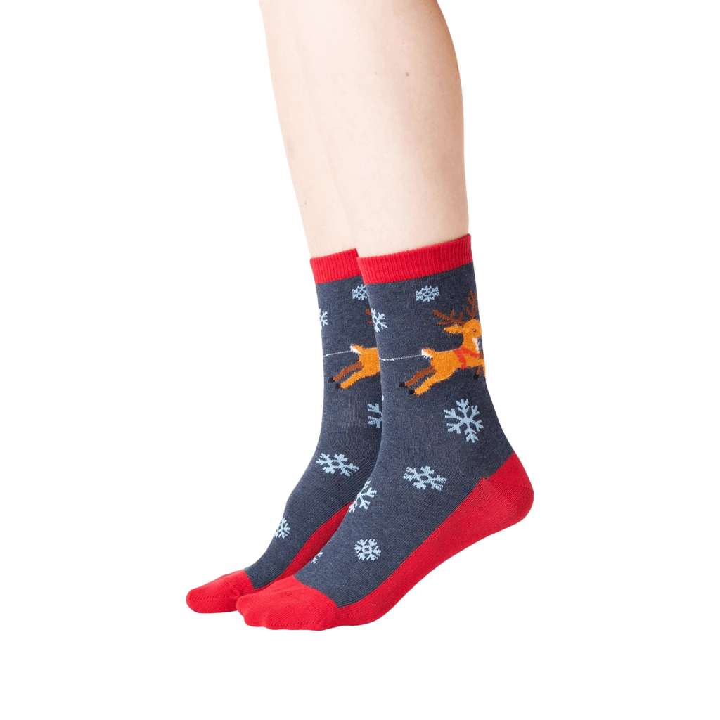 Lifestyle shot showing the socks on a person's feet. The socks are dark gray with red cuffs and toes, featuring a reindeer and snowflake pattern.