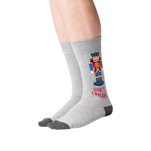 A pair of gray socks with a Nutcracker on them. The Nutcracker is wearing a red and blue outfit and is standing on a brown base. The socks have the words 