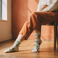 Model wearing cream crew sock with green potted plant pattern, terracotta pants, sitting on wood floor. [lf]
