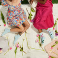 Lifestyle shot of two women wearing Orchid socks, sitting on a blanket surrounded by flowers. [lf]