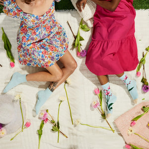 Two women are shown seated on a white, crocheted, picnic blanket, with their legs crossed at the ankles. The woman on the left is wearing a blue floral dress and pale blue socks with a floral pattern. The woman on the right is wearing a pink dress and socks with a floral pattern in shades of pink, purple, and green. They are surrounded by a variety of flowers.