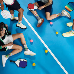 Four people are playing pickleball on a blue court. They are wearing colorful clothing and sneakers. The image is taken from a top-down perspective.