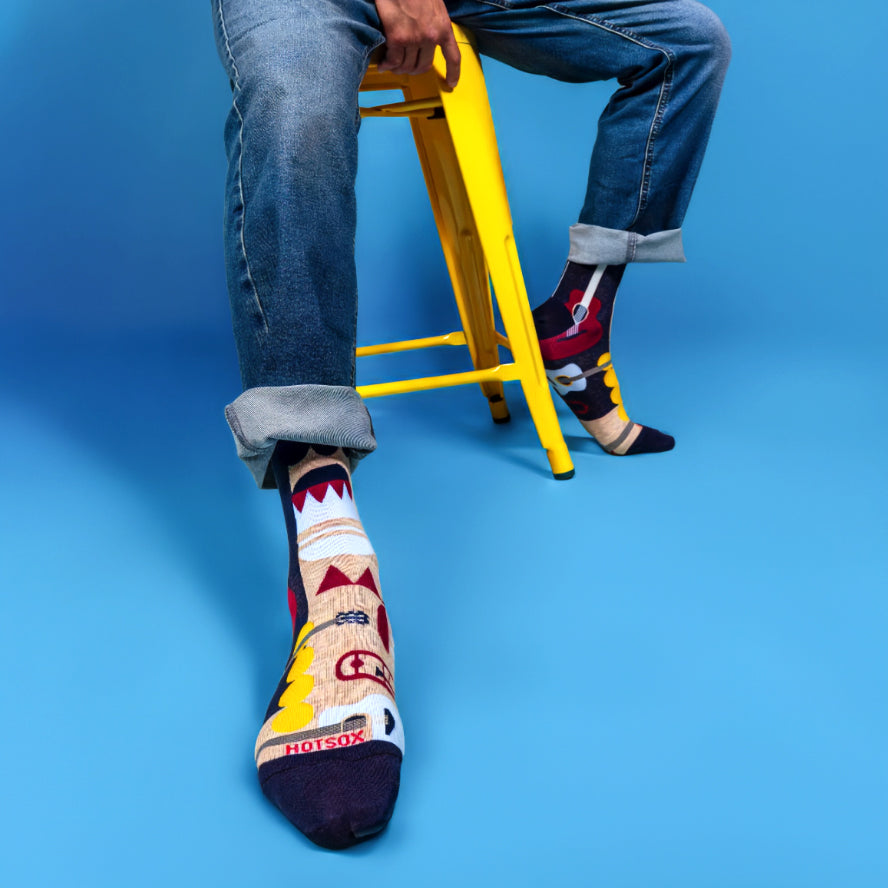 Lifestyle shot of a person wearing the Music Retro socks with jeans, sitting on a yellow stool against a blue background. [lf]