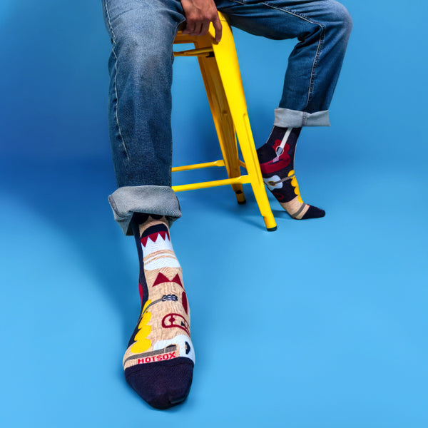 Lifestyle shot of a person wearing the Music Retro socks with jeans, sitting on a yellow stool against a blue background. [lf]
