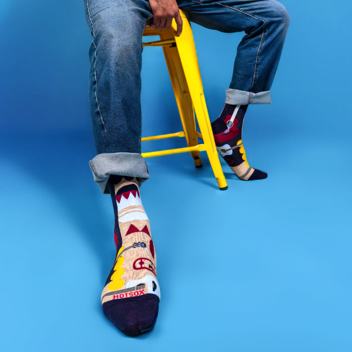 A man is sitting on a yellow stool wearing blue jeans and two different patterned socks. The left sock is dark blue with yellow and red geometric shapes and the word 