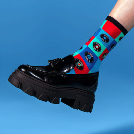 A person's foot is shown from the ankle down. They are wearing a black loafer with a lug sole and bright multi-colored socks with a pattern of red, blue, and green squares. Each square contains a black record.
