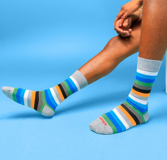 A person is sitting with their legs crossed with one foot resting on the other knee. The person is wearing gray shorts, a white shirt, and colorful striped socks. The socks are blue, green, orange, black, and white.