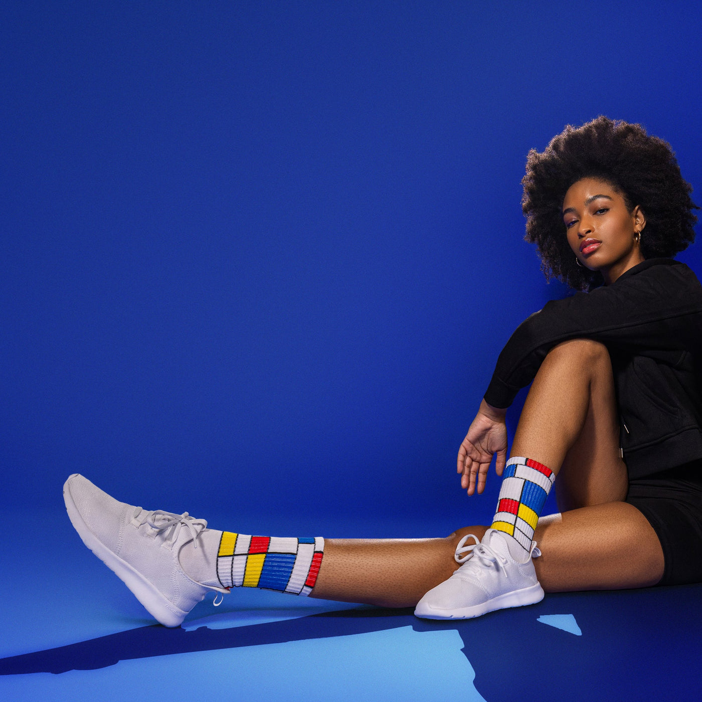 Lifestyle shot of a person wearing Mondrian Active socks with white sneakers against a blue background. [lf]