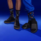 Lifestyle shot showing two people wearing different patterned socks; one is wearing the Art Faces socks with black shoes, the other is wearing orange socks with black slides. Both are shown against a blue background. [lf]