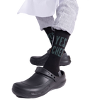 Person wearing "YES, CHEF" black socks with black chef clogs and striped pants, seated on a light wooden block.