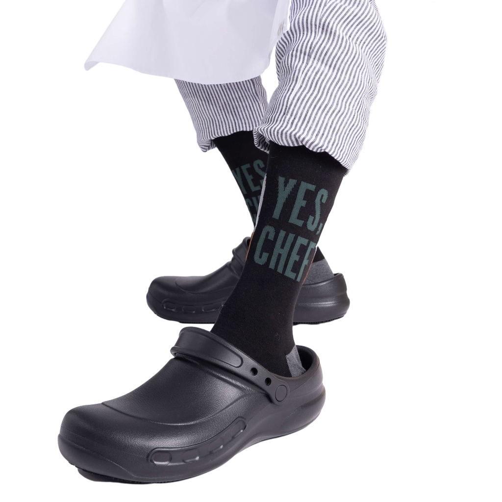 Person wearing "YES, CHEF" black socks with black chef clogs and striped pants, seated on a light wooden block.