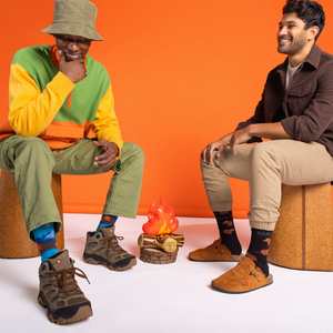 Two men sitting, one wearing 'Dam It' beaver socks with a brown shirt, next to a faux campfire against an orange wall.