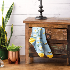 Bee and daisy socks draped over a rustic wooden nightstand with a lamp, snake plant, and copper-potted plant.