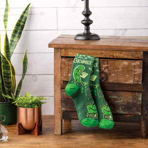 Pair of 'Cool As A Cucumber' socks hanging on a rustic wooden cabinet, next to green potted plants.