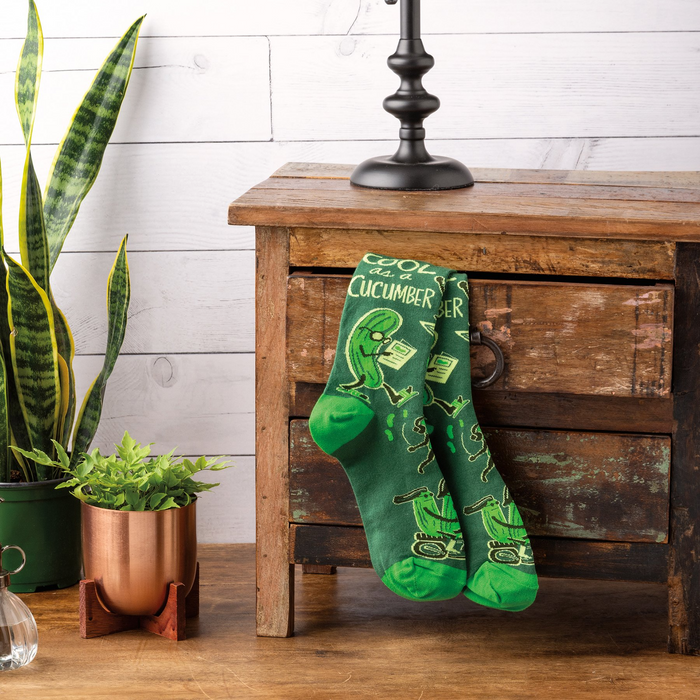 Pair of 'Cool As A Cucumber' socks hanging on a rustic wooden cabinet, next to green potted plants.