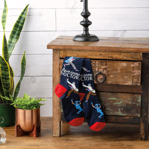 A pair of blue socks with a red toe, heel, and cuff. The socks have a repeating pattern of cartoon doctors in white coats and surgical masks. The socks are packaged on a cardboard display card with a plastic hanger tab. The card has a white background with a red stripe at the top and a blue stripe at the bottom. The words 