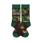 Pair of dark brown crew socks with green cuffs, heels, and toes. Pattern includes trees, deer, and hunters with rifles and bows. Text reads 'I'D RATHER BE HUNTING'.