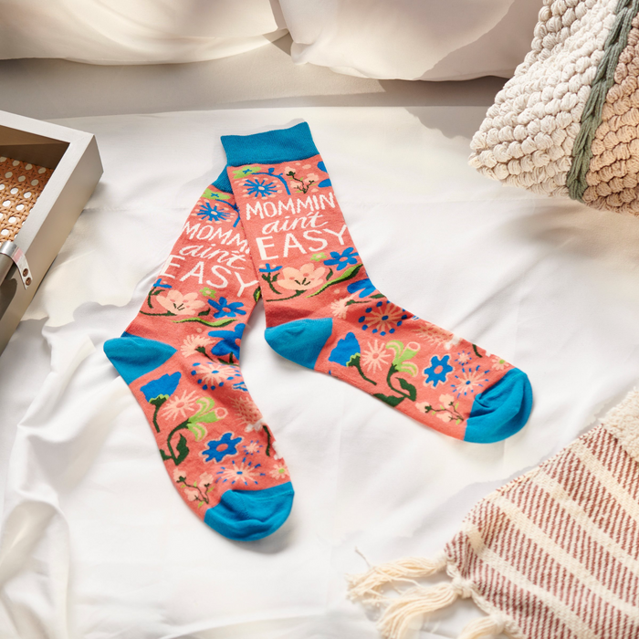 A pair of coral pink crew socks with blue accents and a floral pattern lying on white bedsheets with a woven blanket.