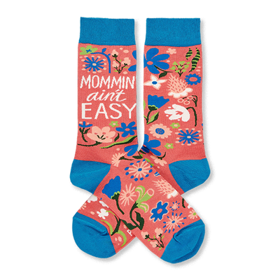 Two coral pink crew socks with blue cuffs, heels, and toes, featuring a colorful floral pattern and "MOMMIN' AIN'T EASY" text.