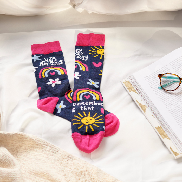 Pair of dark blue socks with pink accents, featuring suns, rainbows, and positive text, laid on white rumpled bedding near an open book and glasses.