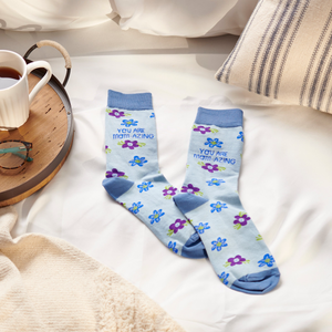 Two light blue floral crew socks laid flat on a white bedspread next to a tea mug and glasses on a tray.