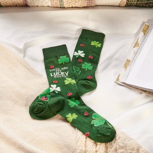 A pair of dark green crew socks with lucky clovers and ladybugs rests on a white bedsheet next to a beige throw and open book.