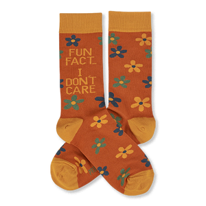 Pair of crew socks in rust orange with yellow cuffs, heels, and toes. Features scattered yellow, teal, and blue flowers. One sock reads 'FUN FACT... I DON'T CARE' in blue and teal text.