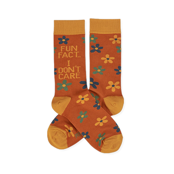 Pair of crew socks in rust orange with yellow cuffs, heels, and toes. Features scattered yellow, teal, and blue flowers. One sock reads 'FUN FACT... I DON'T CARE' in blue and teal text.