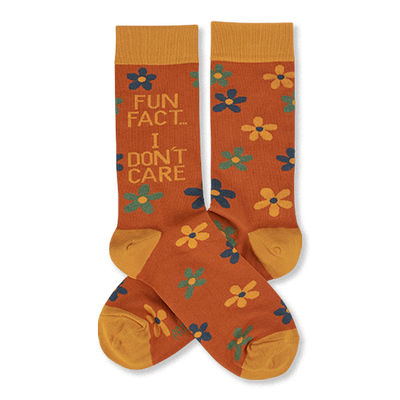 Pair of crew socks in rust orange with yellow cuffs, heels, and toes. Features scattered yellow, teal, and blue flowers. One sock reads 'FUN FACT... I DON'T CARE' in blue and teal text.