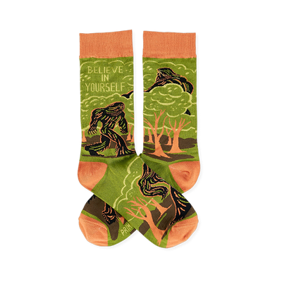A pair of crew socks, olive green with orange accents. Features a dark green Bigfoot-like creature in a forest and 'Believe In Yourself' text.