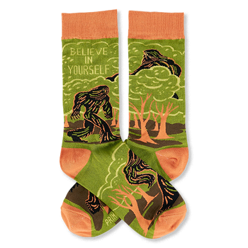 A pair of crew socks, olive green with orange accents. Features a dark green Bigfoot-like creature in a forest and 'Believe In Yourself' text.