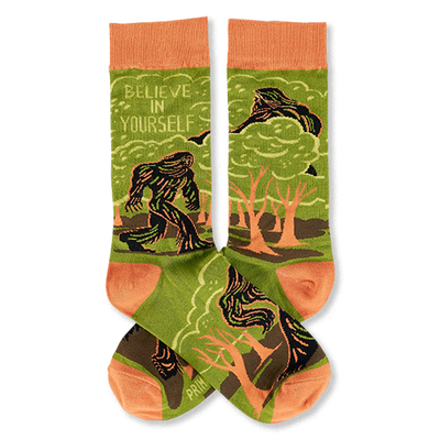 A pair of crew socks, olive green with orange accents. Features a dark green Bigfoot-like creature in a forest and 'Believe In Yourself' text.