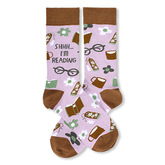 A pair of light purple crew socks with a brown cuff, toe, and heel, featuring a whimsical reading-themed pattern of books, coffee cups, eyeglasses, and the text 'SHHH.