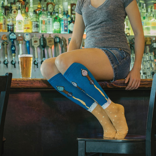 A woman perches on a bar top in cutoff denim shorts beside a pint of golden lager, rows of beer tap handles and liquor bottles behind her, wearing the Beer Taps socks in BLUE FOG with illustrated tap handles and frothy beer pouring into a tan foot. [lf]