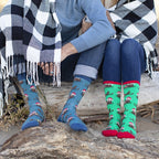 Couple sitting on driftwood at the beach wrapped in a black and white buffalo-check blanket, their feet side by side in Otterly Merry socks, one pair blue with red Santa-hat otters and the other green with the same festive otter print and red toes. [lf]
