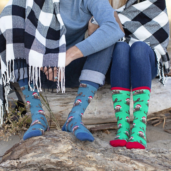 Couple sitting on driftwood at the beach wrapped in a black and white buffalo-check blanket, their feet side by side in Otterly Merry socks, one pair blue with red Santa-hat otters and the other green with the same festive otter print and red toes. [lf]
