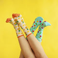 Two pairs of upside-down legs cross at the ankles against a bright yellow backdrop, one foot in cream Partial To Pineapples socks with orange cuffs and the other in turquoise cherry-print socks. [lf]