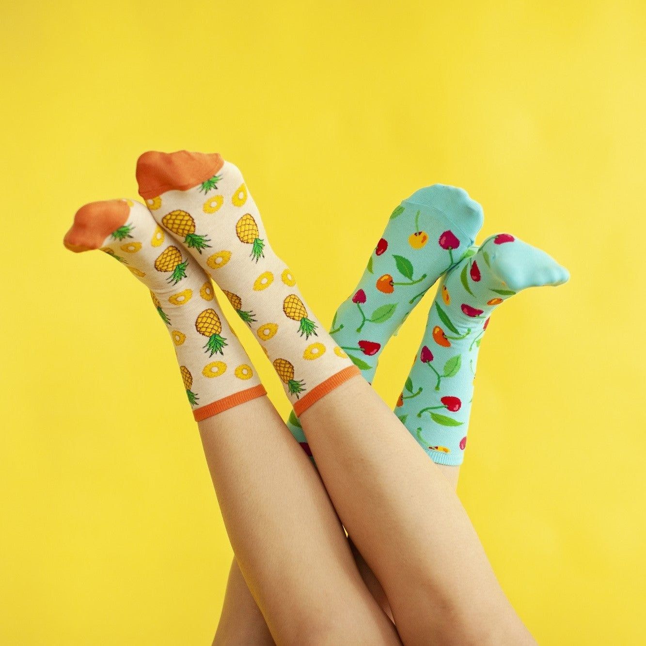 Two pairs of upside-down legs cross at the ankles against a bright yellow backdrop, one foot in cream Partial To Pineapples socks with orange cuffs and the other in turquoise cherry-print socks. [lf]