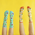 Two pairs of legs point skyward in front of a sunny yellow wall, showing off Partial To Pineapples socks beside a turquoise cherry pair with leafy stems. [lf]
