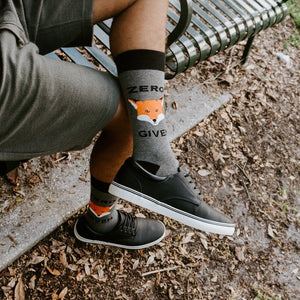 Cropped view of legs seated on a metal park bench, ZERO and GIVE wording wrapping the cuffs of Zero Fox Given socks above black sneakers. [lf]