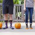 A couple stands shoulder to shoulder on a wood porch flanked by three orange pumpkins, the man in shorts showing the Jack O' All Trades socks in navy printed with grinning orange jack-o-lanterns alongside his partner's candy-corn pair. [lf]