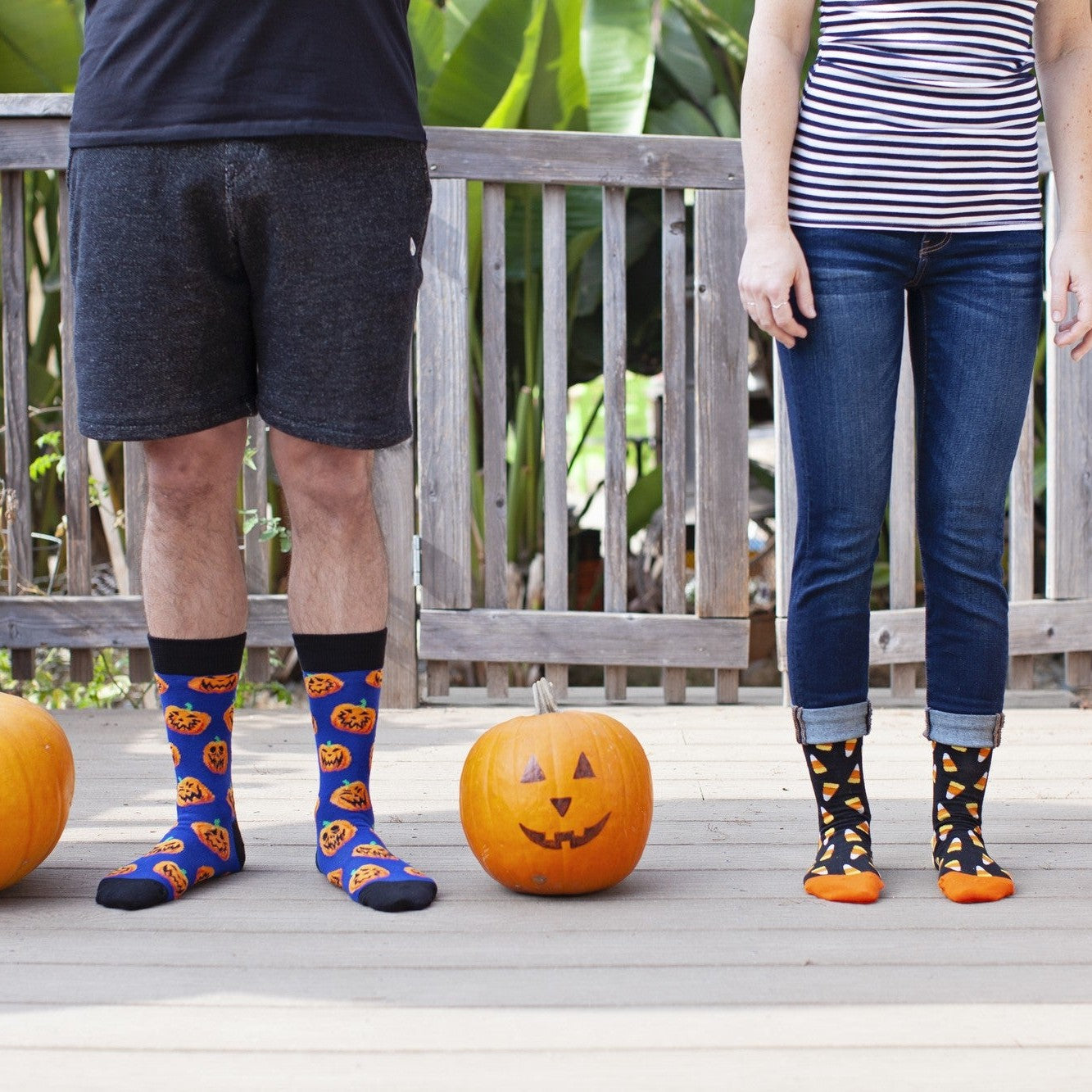A couple stands shoulder to shoulder on a wood porch flanked by three orange pumpkins, the man in shorts showing the Jack O' All Trades socks in navy printed with grinning orange jack-o-lanterns alongside his partner's candy-corn pair. [lf]