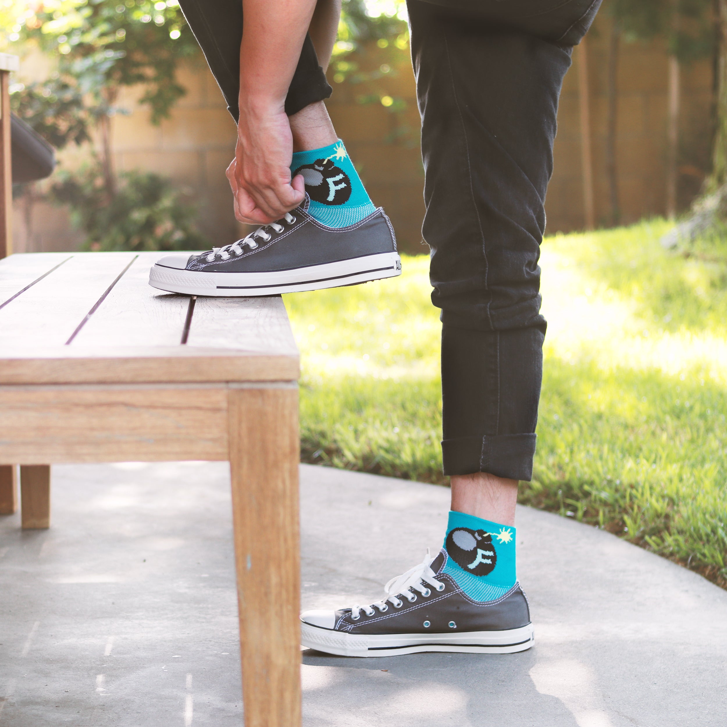 Lifestyle shot: Person putting on teal 'F'Bomb socks with black Converse sneakers. [lf]