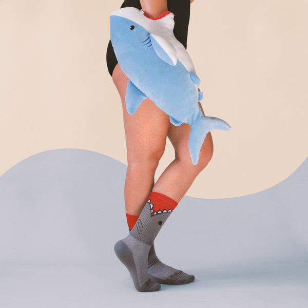 Lifestyle photo showing a person wearing the 'Bite Me' shark socks with a plush shark toy. [lf]