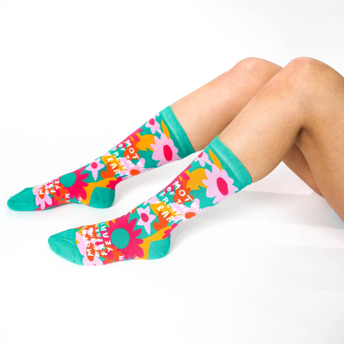 A person's legs, bent at the knees, wearing the green, pink, orange, and yellow floral 'Mother Of The Freakin' Year' crew socks on a white background.