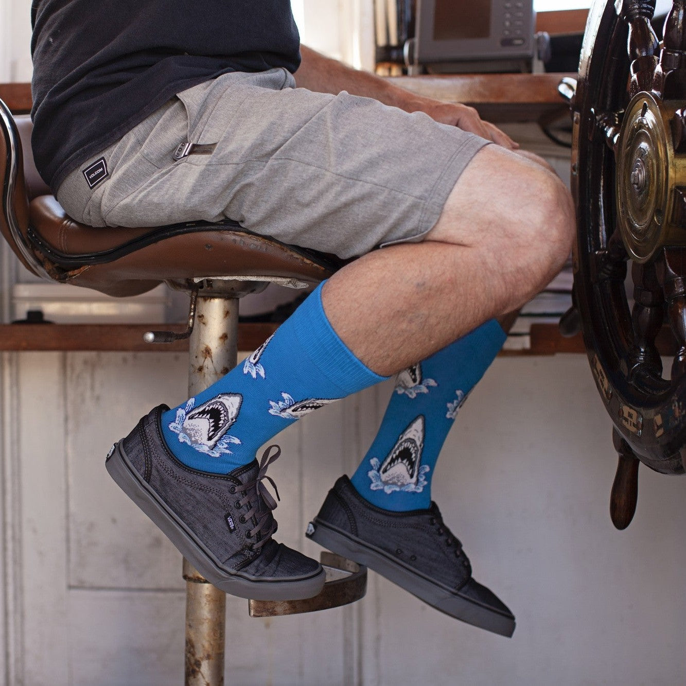 Side angle of crossed legs on a boat's leather pilot seat near the wheel, highlighting Shark Attack XL in BLUE socks with shark and splash motifs above black canvas sneakers. [lf]