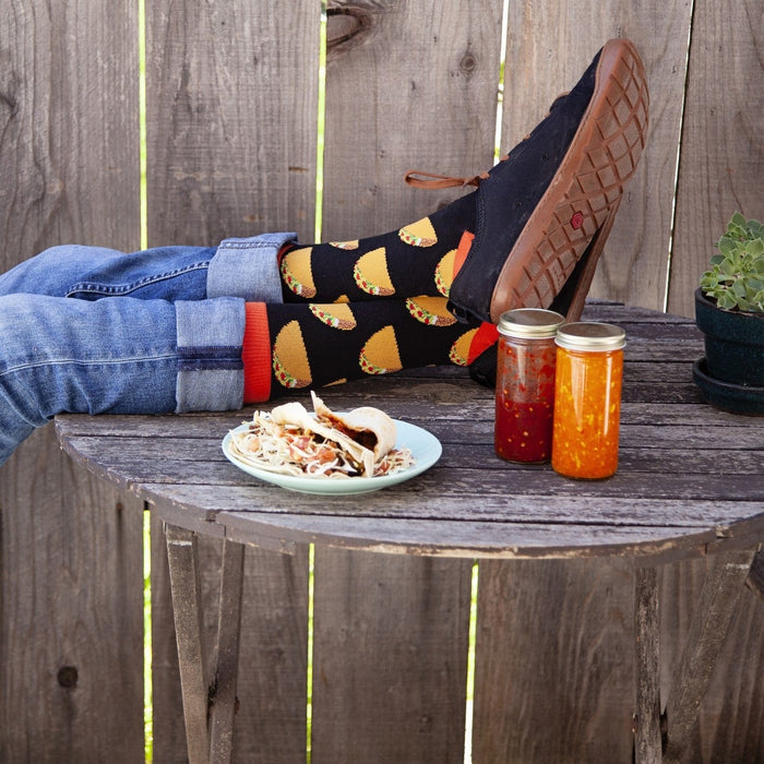 A man kicks back on a wooden table with a beer in hand, a plate of tacos and jars of salsa beside him, wearing brown leather sandals over the black Tacos XL socks printed with hand-drawn taco illustrations. [lf]