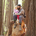 A bearded dad in a plaid flannel and his daughter in a pink-striped hoodie sit on a giant redwood stump, both wearing Big Foot Christmas socks tucked into hiking boots as sunlight filters through the trees. [lf]