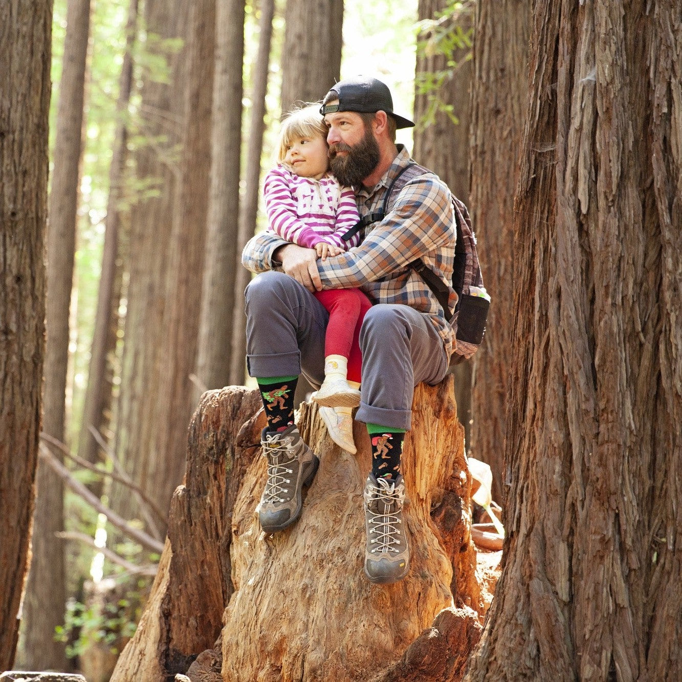 A bearded dad in a plaid flannel and his daughter in a pink-striped hoodie sit on a giant redwood stump, both wearing Big Foot Christmas socks tucked into hiking boots as sunlight filters through the trees. [lf]